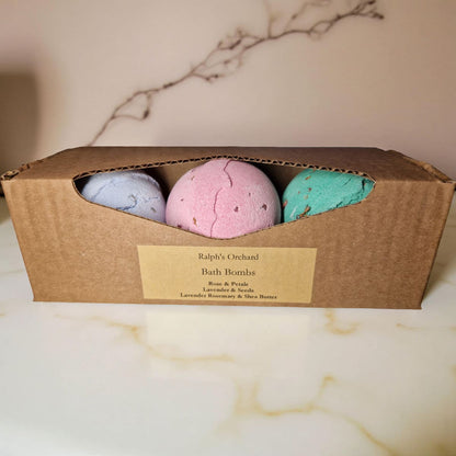 Three bath bombs in a cardboard box labeled 'Ralph's Orchard' on a marble surface.