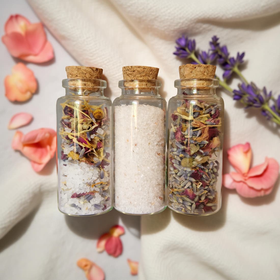 Three small glass jars with cork lids containing dried flowers and herbs on a soft white background with pink and purple flowers.