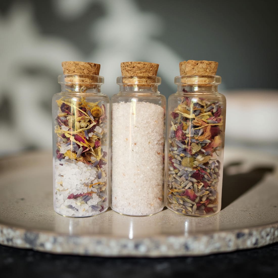 Three glass jars with cork lids containing various herbs and spices on a stone surface.
