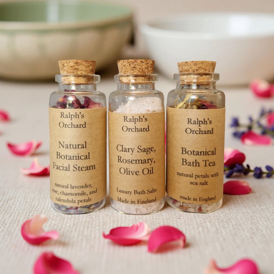 Three glass bottles labeled 'Ralph's Orchard' with cork lids on a surface with pink petals.