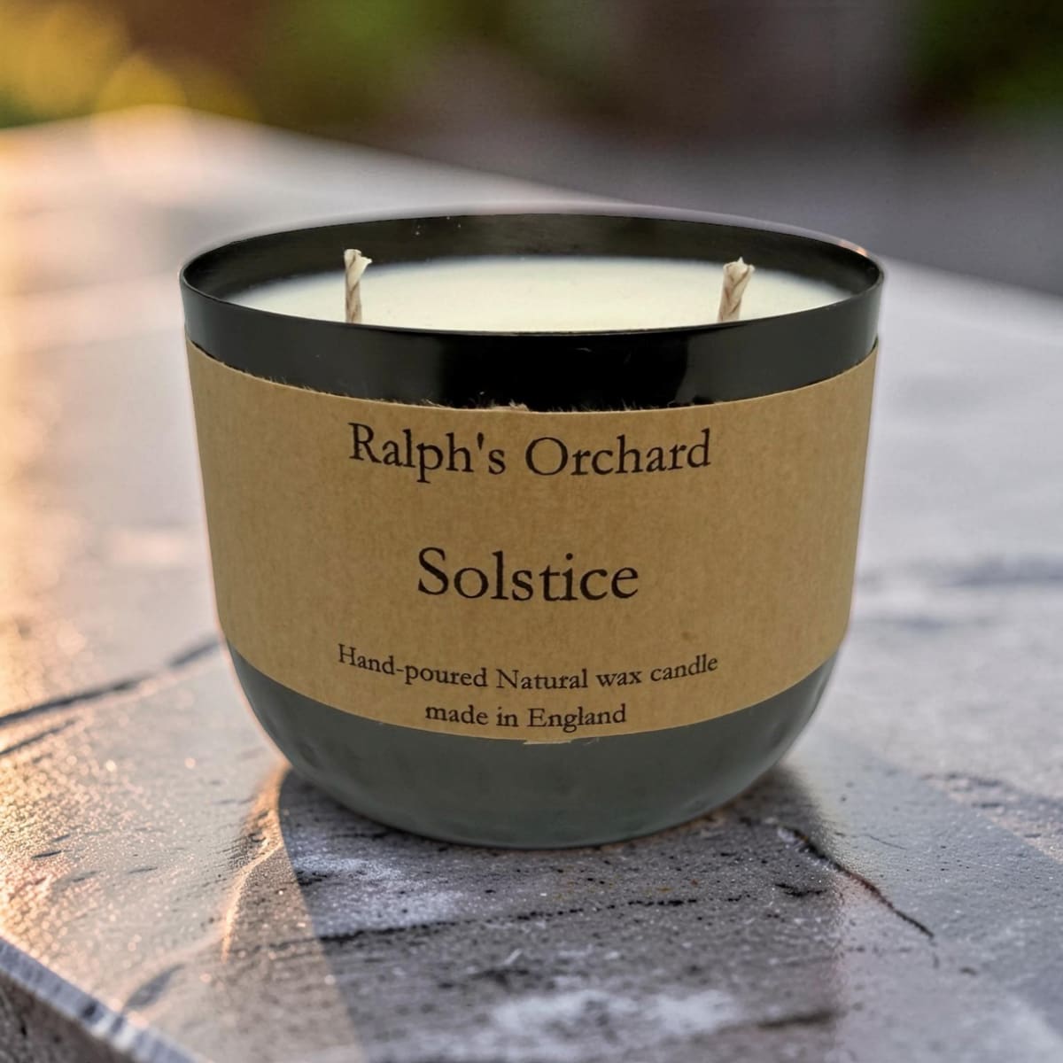 A two-wick hand-poured natural wax candle in a dark gray glass jar with a black rim and a brown kraft paper label reading 'Ralph's Orchard Solstice'. The candle is placed on a textured gray stone surface with a blurred outdoor background.