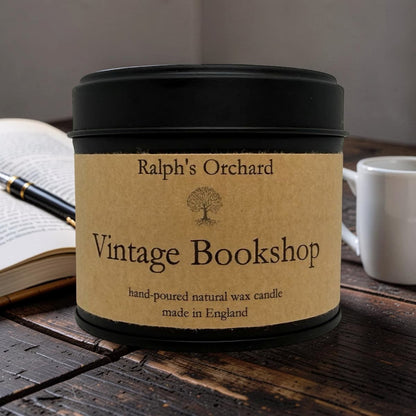 A black metal candle tin with a kraft paper label reading 'Ralph's Orchard Vintage Bookshop' sits on a rustic wooden table, styled with an open book, pen, and white mug in the background.