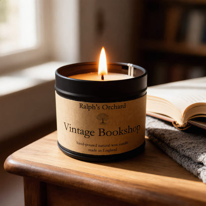 A lit candle in a black metal tin with a tan paper label reading 'Ralph's Orchard Vintage Bookshop,' placed on a wooden table beside an open book and a folded gray knit blanket in a softly lit indoor setting.