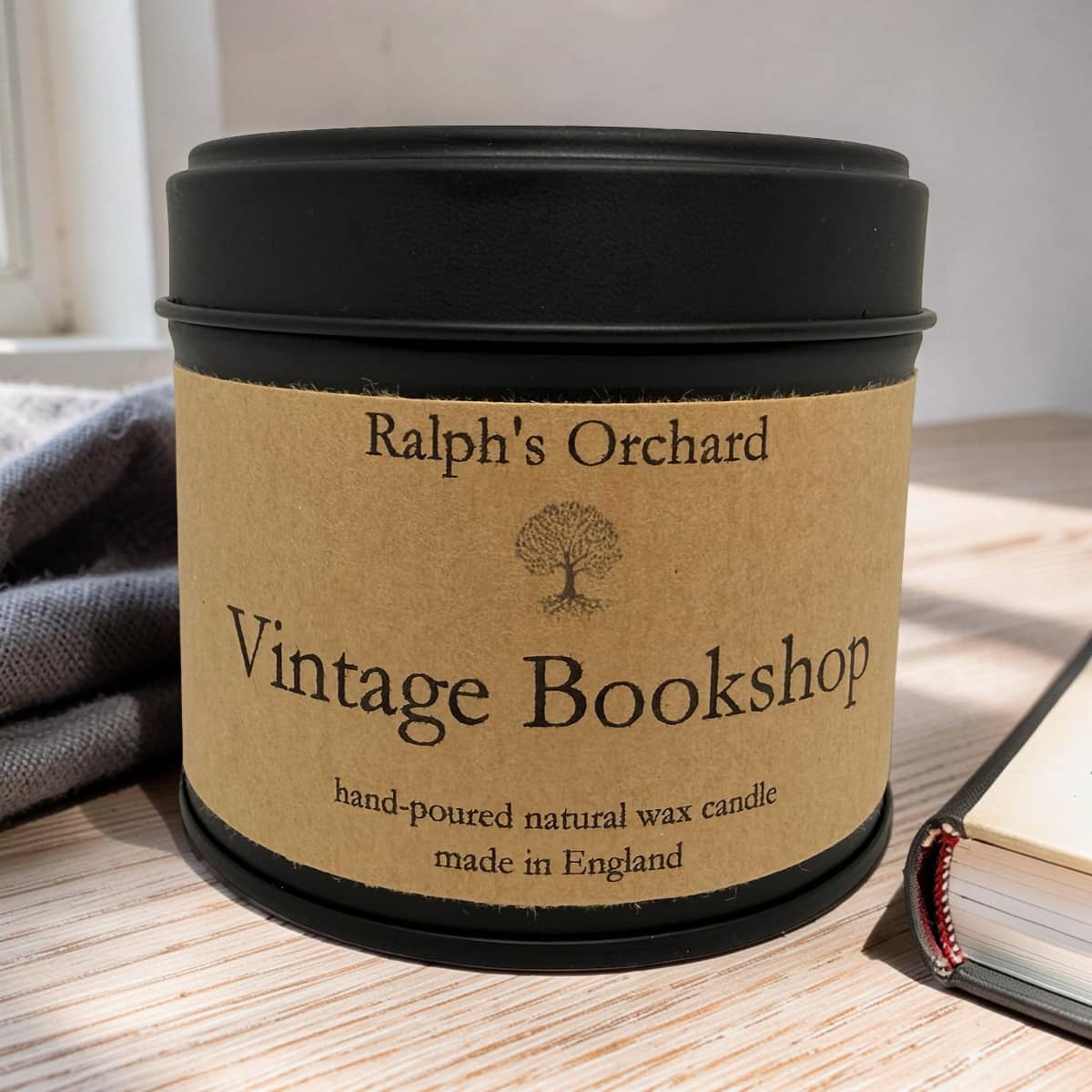 A black metal candle tin with a kraft paper label reading 'Ralph's Orchard Vintage Bookshop' sits on a wooden tabletop. A folded gray cloth is on the left, and part of a book is visible on the right.