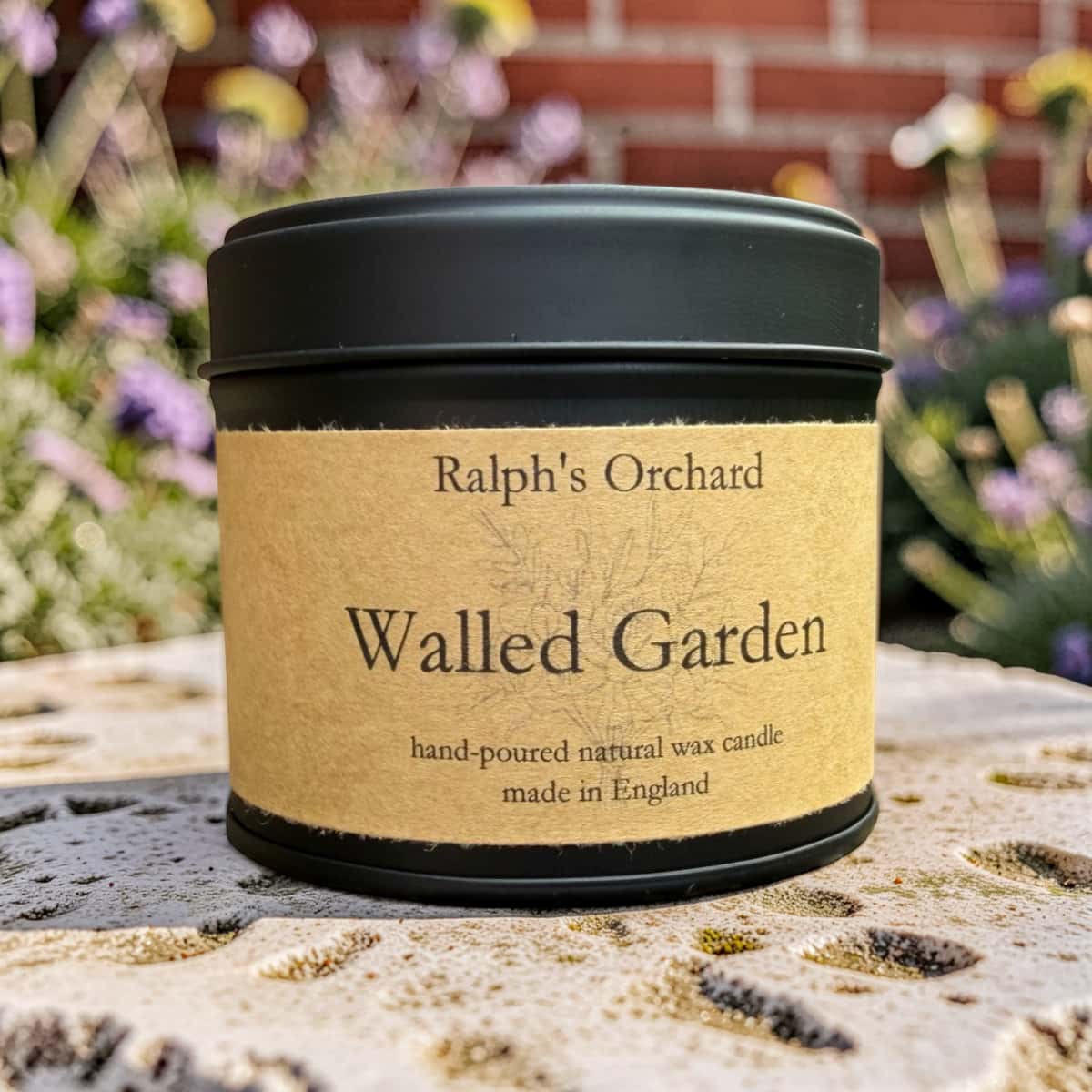 A black metal candle tin with a kraft paper label reading 'Ralph's Orchard' and 'Walled Garden', photographed outdoors on a stone surface with blurred flowers and a brick wall in the background.