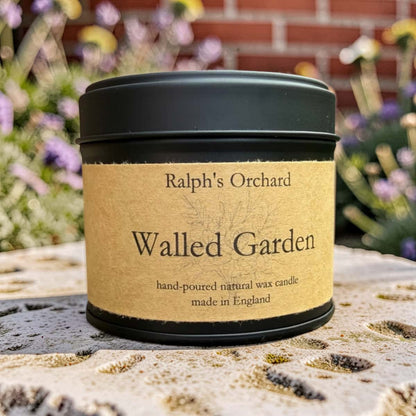 A black metal candle tin with a kraft paper label reading 'Ralph's Orchard' and 'Walled Garden', photographed outdoors on a stone surface with blurred flowers and a brick wall in the background.