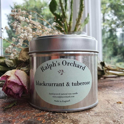 Candle tin labeled 'Ralph's Orchard blackcurrant & tuberose' on a wooden surface with flowers in the background.