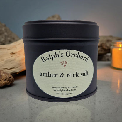 Candle tin labeled 'Ralph's Orchard amber & rock salt' on a textured surface with rocks and a candle in the background.