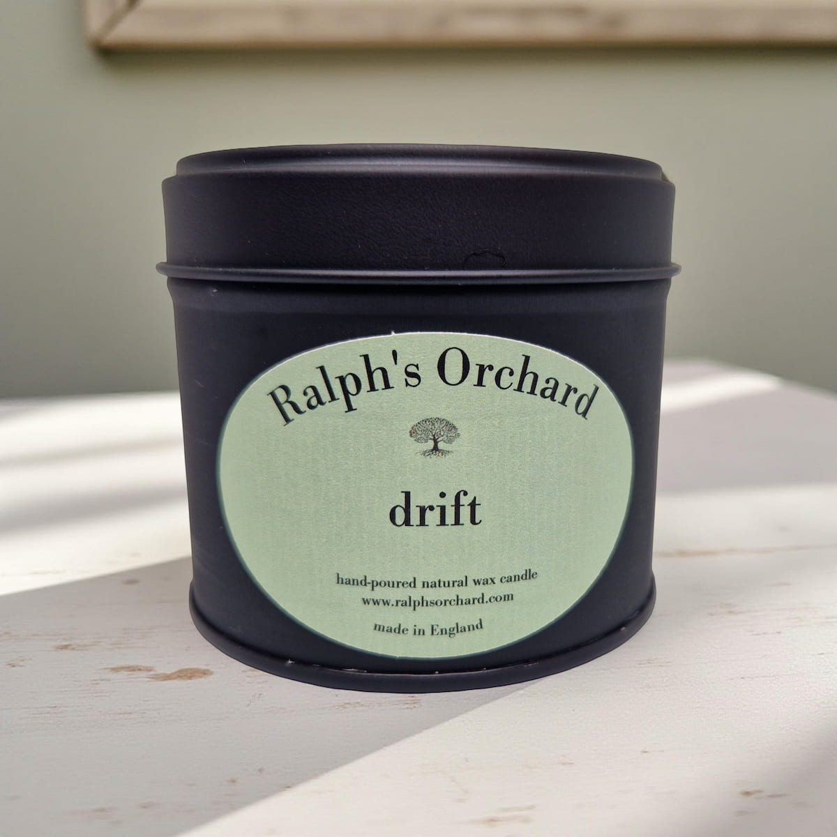 Candle in a black tin with 'Ralph's Orchard' label on a light wooden surface.