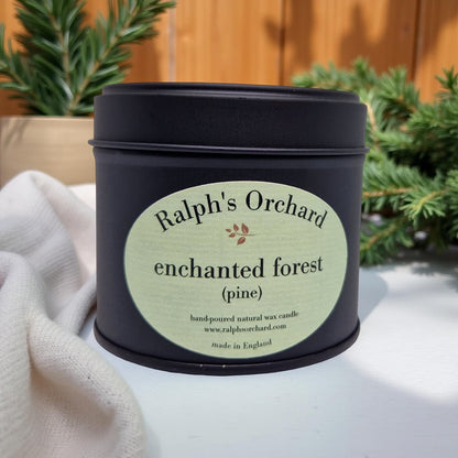 Candle tin labeled 'Ralph's Orchard Enchanted Forest Pine' on a light surface with plants in the background.