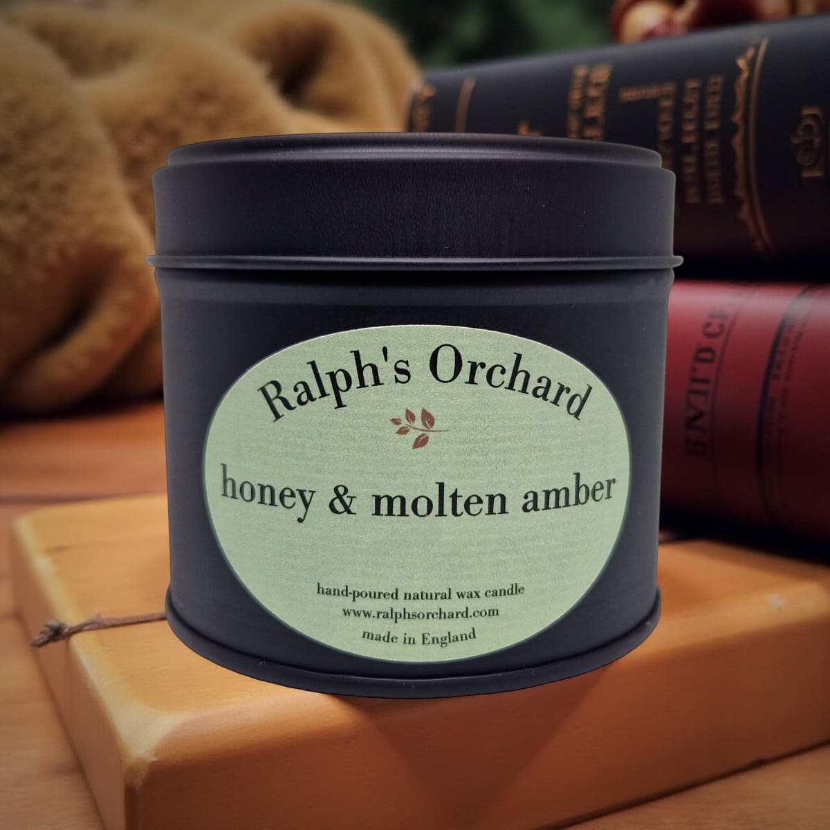 Candle tin labeled 'Ralph's Orchard honey & molten amber' on a wooden surface with books in the background.