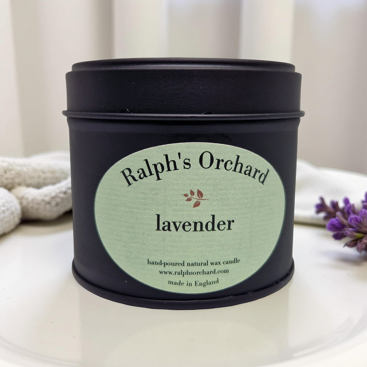 Candle tin labeled 'Ralph's Orchard lavender' on a white surface with a blurred background