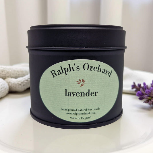 Candle tin labeled 'Ralph's Orchard lavender' on a white surface with a blurred background