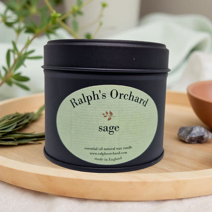 Candle in a black tin labeled 'Ralph's Orchard Sage' on a wooden surface with plants in the background.