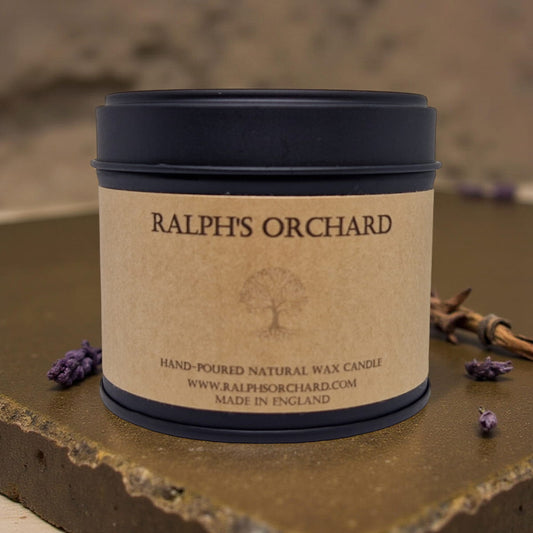 Candle in a tin labeled 'Ralph's Orchard' on a stone surface with lavender flowers.