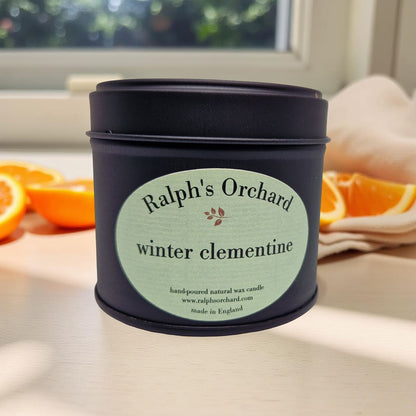 Candle in a black tin labeled 'Ralph's Orchard winter clementine' on a surface with oranges.