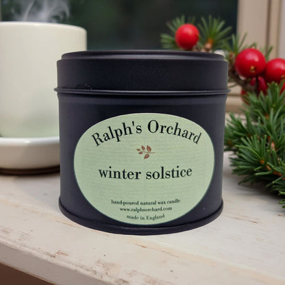 Candle in a black tin labeled 'Ralph's Orchard winter solstice' on a wooden surface with decorative elements.