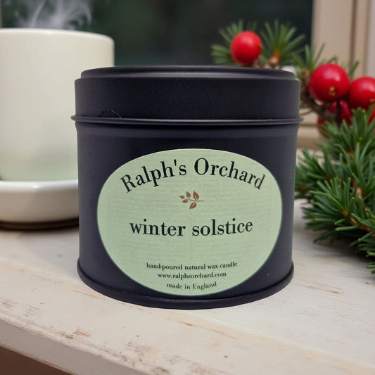Candle in a black tin labeled 'Ralph's Orchard winter solstice' on a wooden surface with decorative elements.