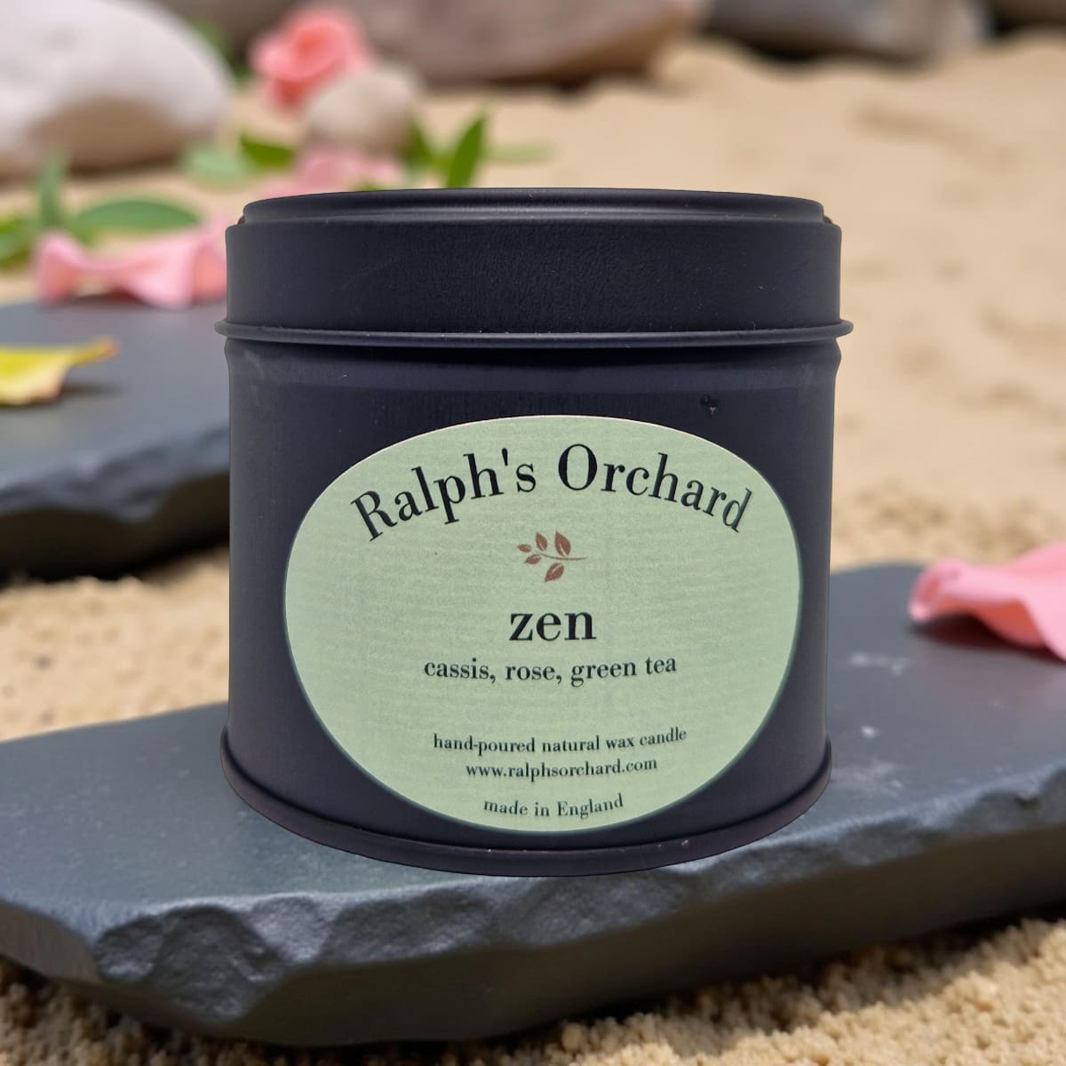 Candle tin labeled 'Ralph's Orchard Zen' on a stone surface with flowers in the background