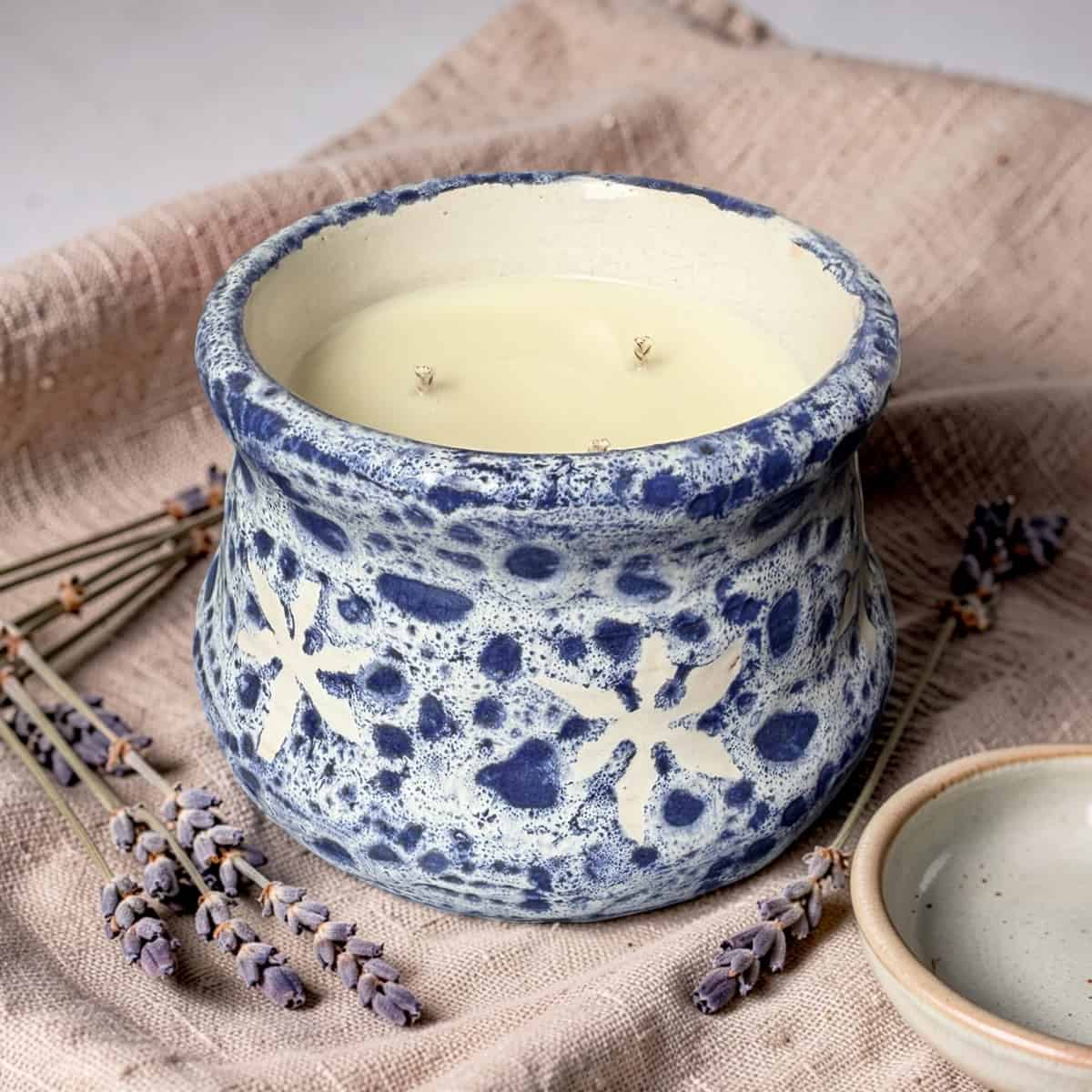 Ceramic candle holder with a candle inside, surrounded by lavender on a textured fabric background