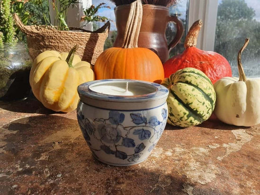 Ceramic container with a lit candle surrounded by pumpkins and gourds on a windowsill.