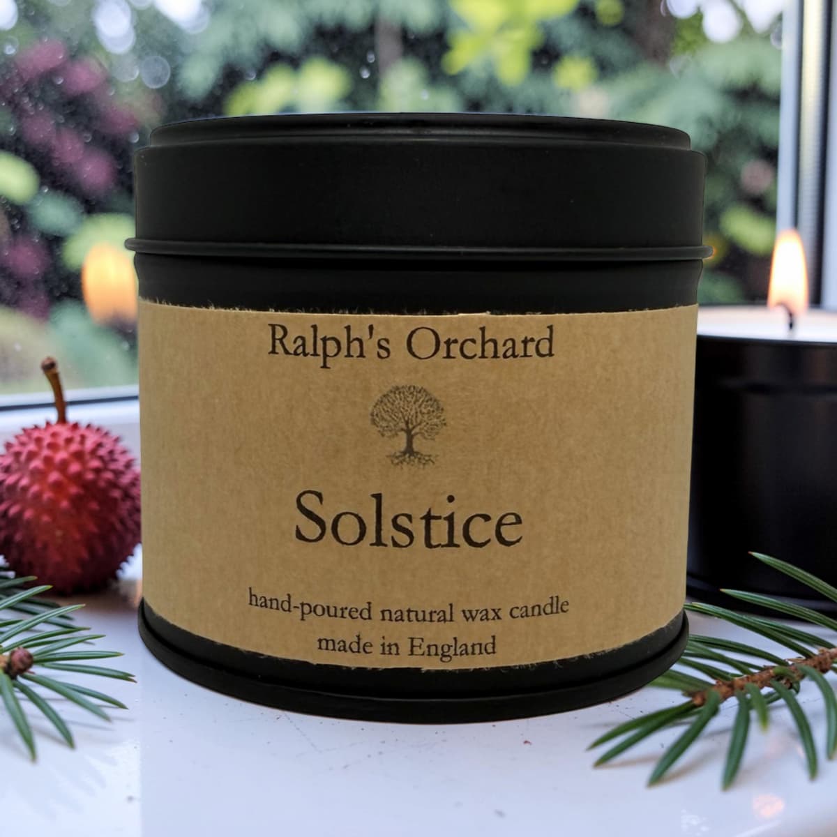 A black metal candle tin with a kraft paper label reading 'Ralph's Orchard Solstice hand-poured natural wax candle made in England,' styled on a white surface with pine branches and a red berry against a softly blurred window background.