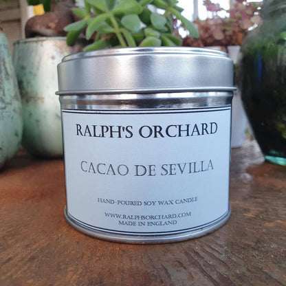 Candle tin labeled 'Ralph's Orchard Cacao de Sevilla' on a wooden surface with plants in the background.