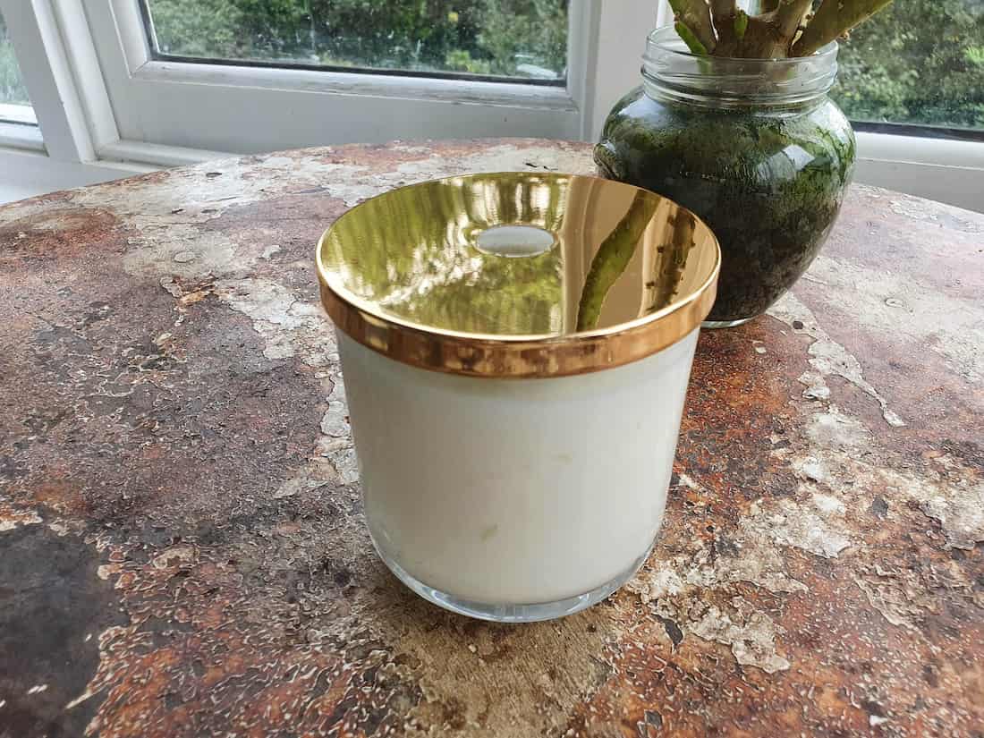 White candle with gold rim on a textured surface, with a jar of greenery in the background.