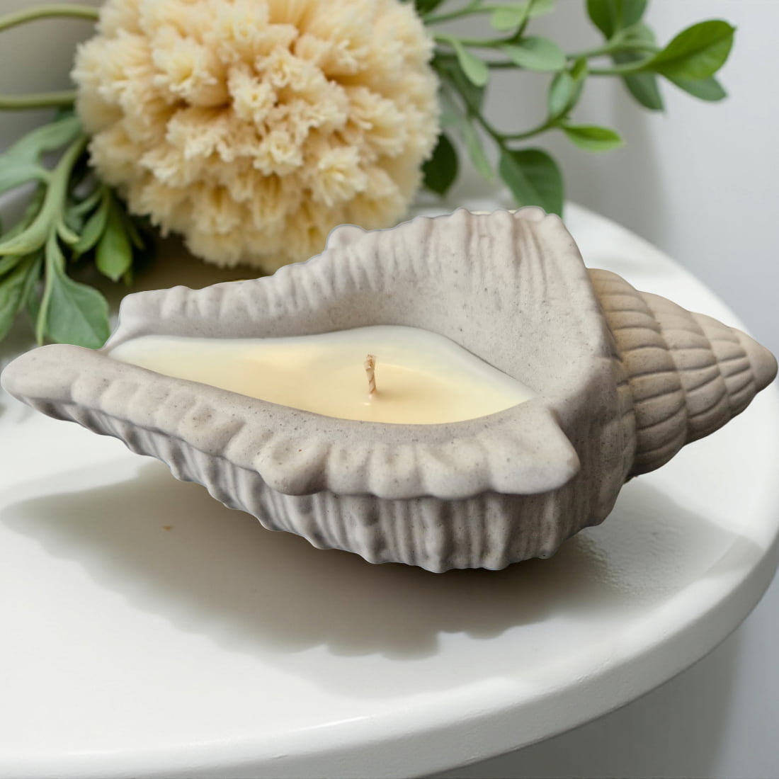 Shell-shaped candle with a small flame on a white surface with flowers and leaves in the background