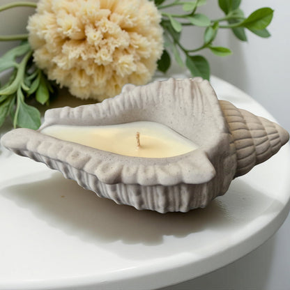 Shell-shaped candle with a small flame on a white surface with flowers and leaves in the background