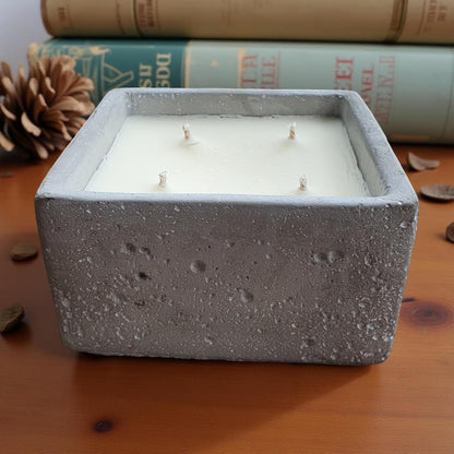 Square concrete candle with 4 wicks on a wooden surface with books and pine cones in the background.