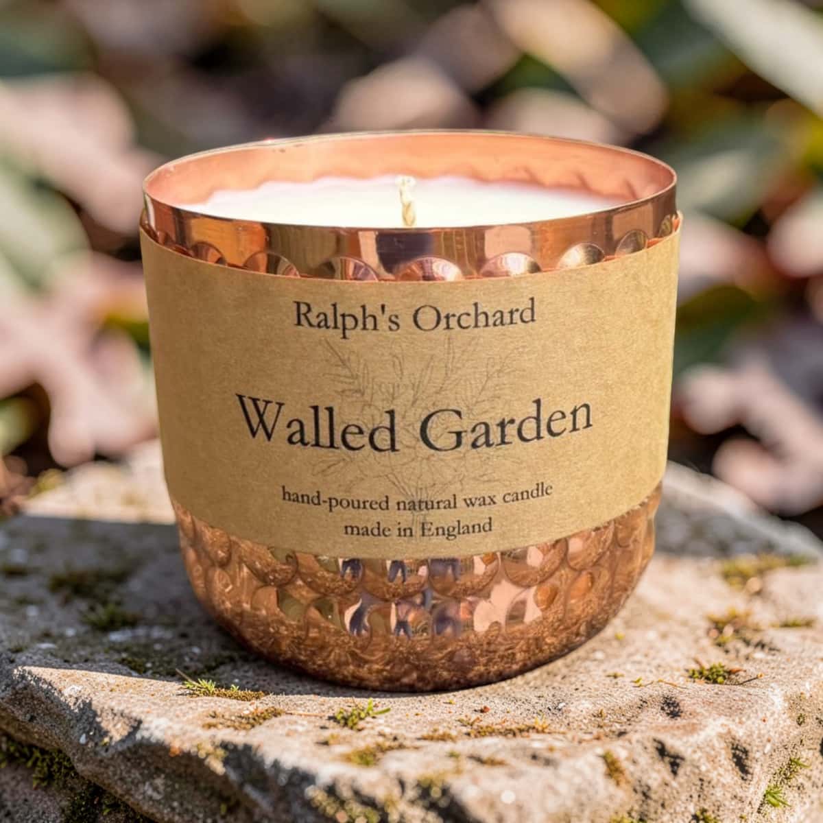 A copper-colored glass candle jar with a hammered finish and a kraft paper label reading 'Ralph's Orchard Walled Garden', photographed outdoors on a stone surface with blurred foliage in the background.