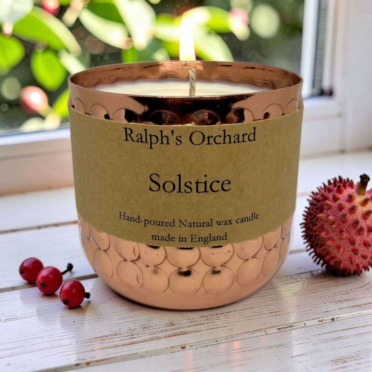 A lit hand-poured natural wax candle in a hammered copper-colored vessel with a kraft paper label reading 'Ralph's Orchard Solstice'. The candle sits on a white wooden surface with red berries and a spiky pink fruit nearby, with a blurred window and greenery in the background.