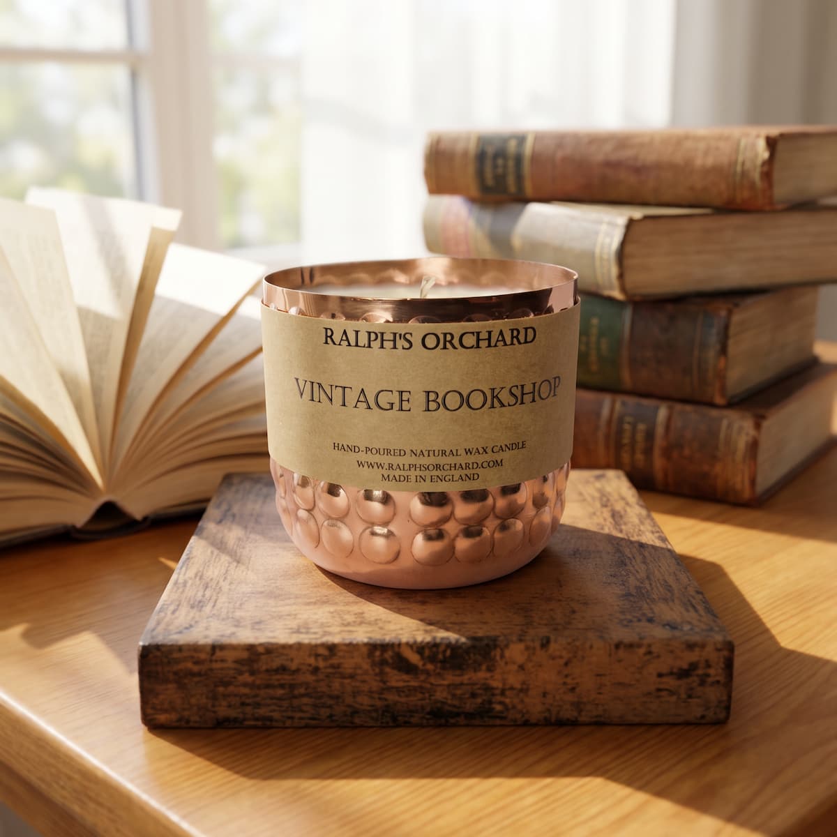 A copper-colored candle vessel with a beige paper label reading 'RALPH'S ORCHARD VINTAGE BOOKSHOP' sits on a wooden block on a wooden table, with an open book and stacked vintage books in a softly lit room background.