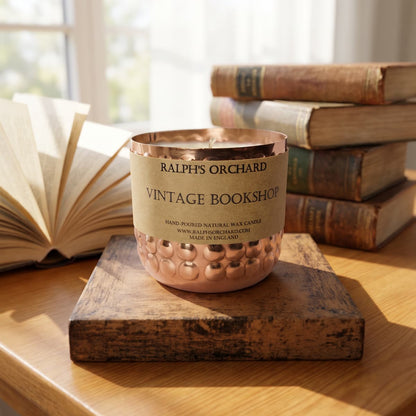 A copper-colored candle vessel with a beige paper label reading 'RALPH'S ORCHARD VINTAGE BOOKSHOP' sits on a wooden block on a wooden table, with an open book and stacked vintage books in a softly lit room background.