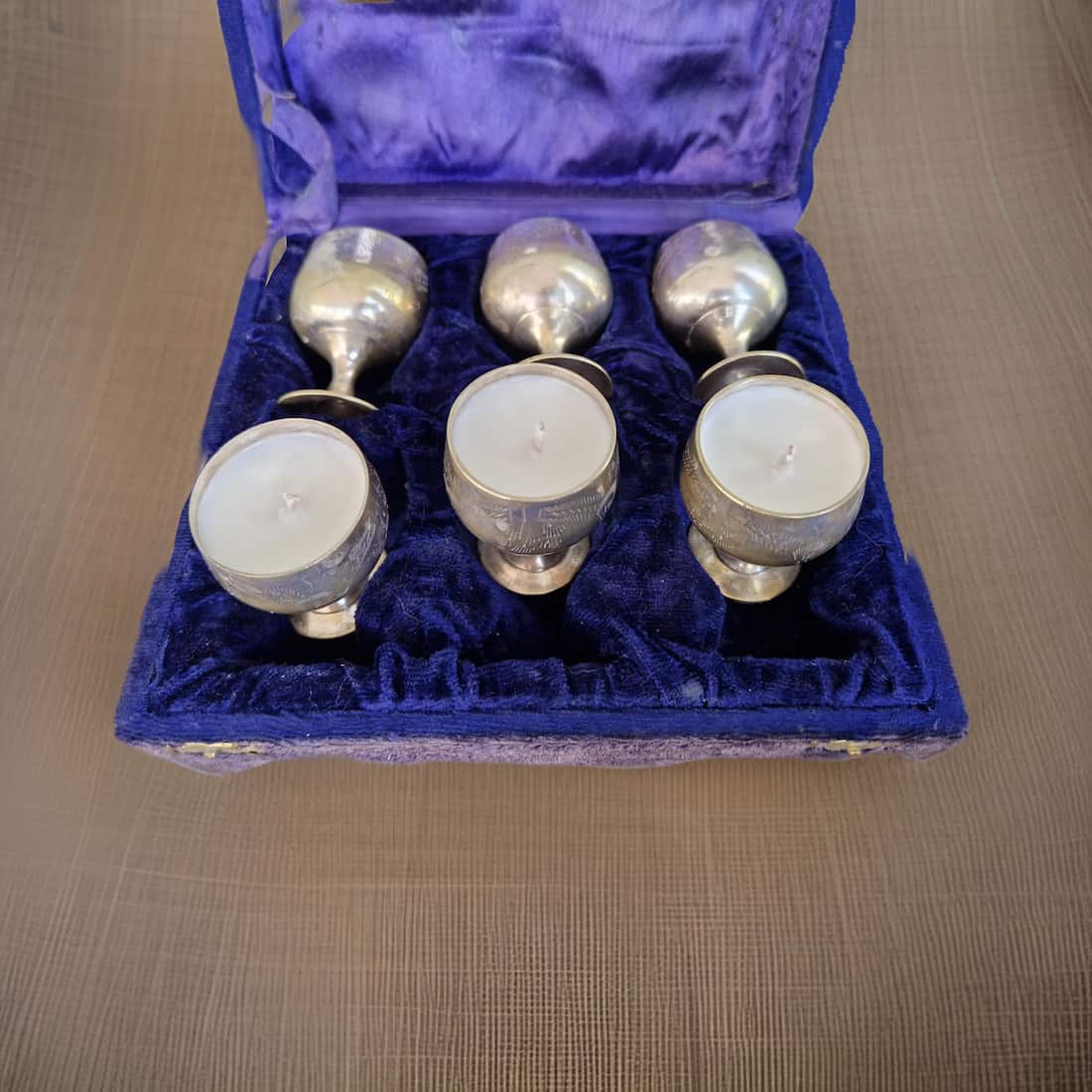 Set of six silver candle holders with white candles in a blue velvet box on a wooden surface.
