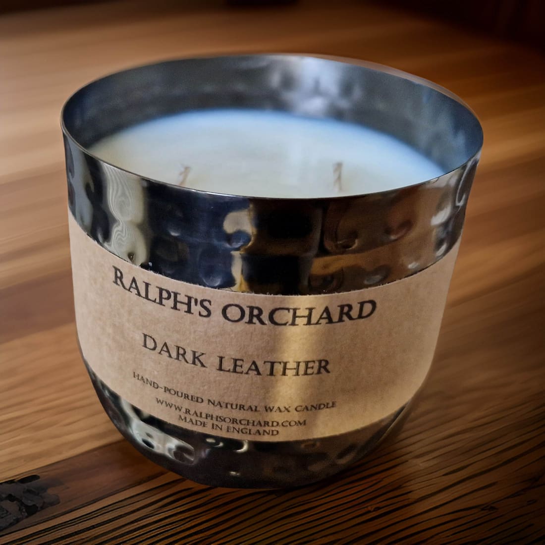 Candle in a metallic container labeled 'Ralph's Orchard Dark Leather' on a wooden surface.