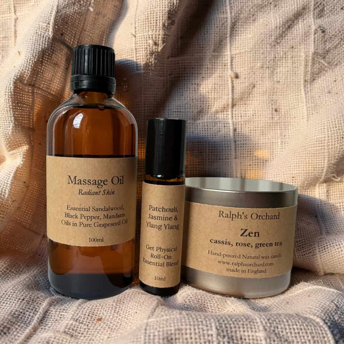 Three wellness products including a massage oil bottle, essential oil roller, and candle on a textured fabric background.