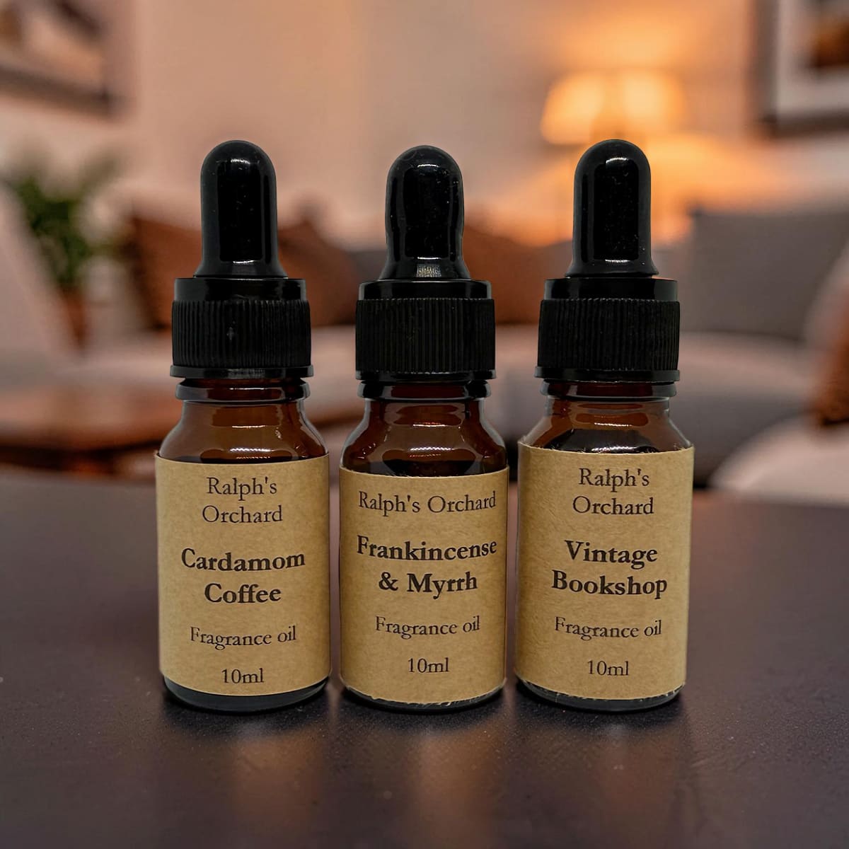 Three bottles of Ralph's Orchard fragrance oils on a wooden surface with a blurred indoor background.