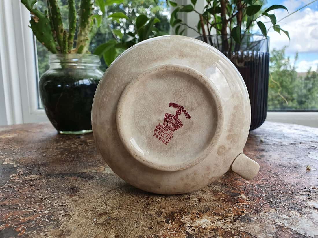 Ceramic teapot with a visible brand logo on a wooden surface with plants in the background
