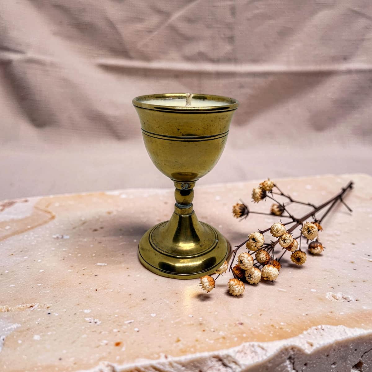 A small brass goblet-shaped candle holder with a wick, styled on a textured stone slab next to dried baby's breath flowers against a draped fabric backdrop.
