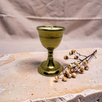 A small brass goblet-shaped candle holder with a wick, styled on a textured stone slab next to dried baby's breath flowers against a draped fabric backdrop.