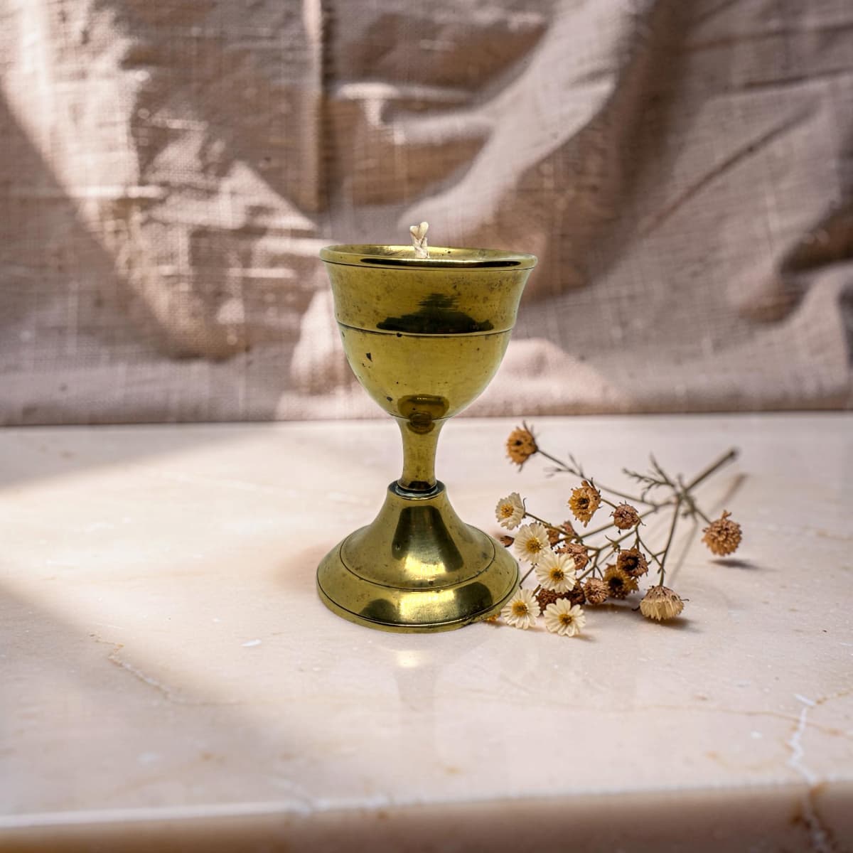 A small polished brass goblet or candle holder with a matching wick, placed on a light stone surface beside dried white flowers against a draped fabric backdrop.