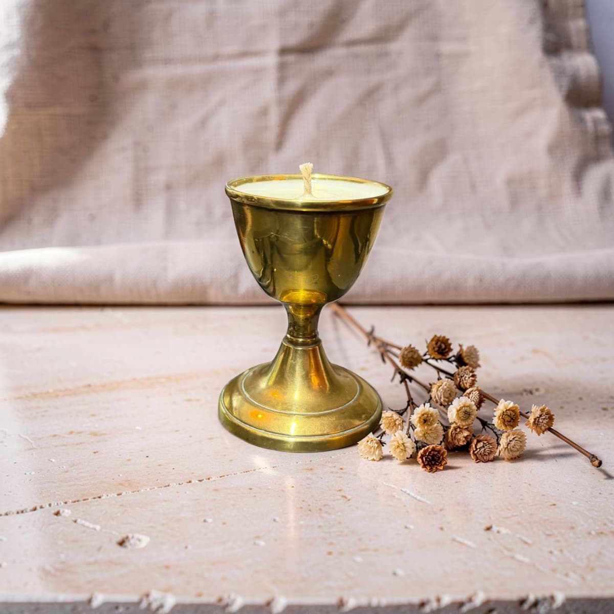 A brass candle holder candle with a small wick, styled on a rustic wooden surface next to dried flowers against a neutral linen background.