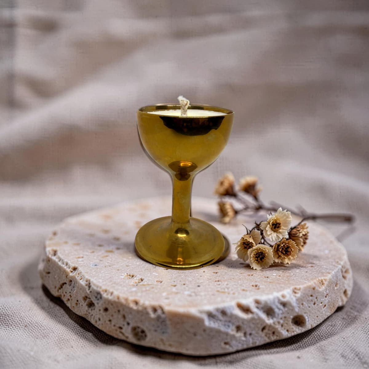 A small gold goblet-shaped candle with a wick sits on a round stone slab, styled with dried flowers against a soft beige fabric backdrop.