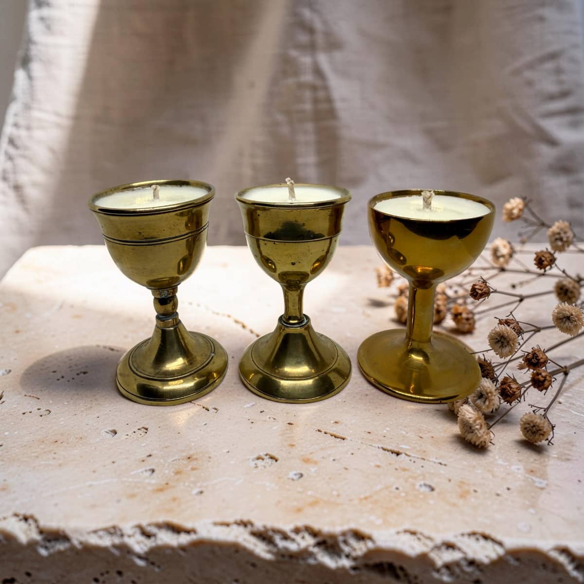 Three brass candle holders with white candles, arranged on a rustic stone surface next to dried flowers against a draped fabric backdrop.