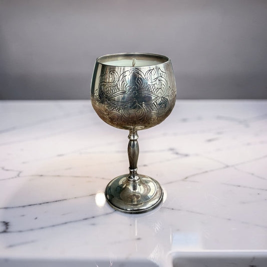 An ornate silver-toned metal goblet or candle holder with engraved floral detailing, standing on a white marble surface against a gray background.