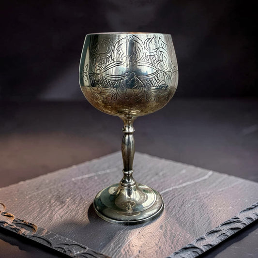 An ornate silver goblet with intricate engraved patterns, resting on a dark slate coaster against a dark, moody background.