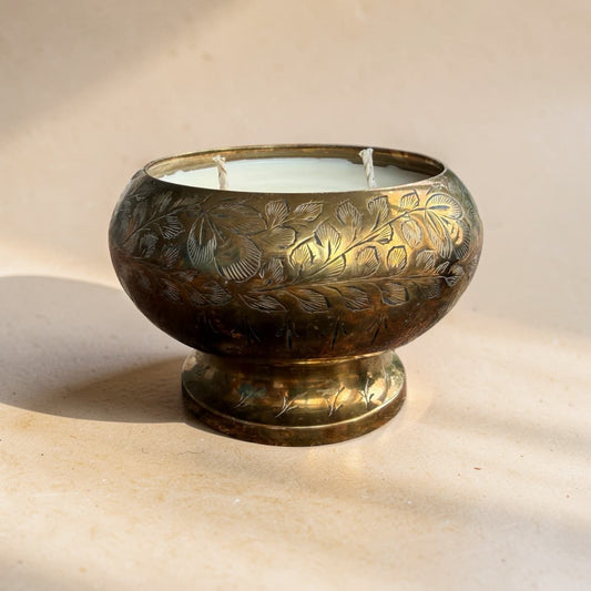 An ornate round brass candle holder with engraved floral patterns, holding a white double-wick candle, set against a warm beige background with soft shadows.