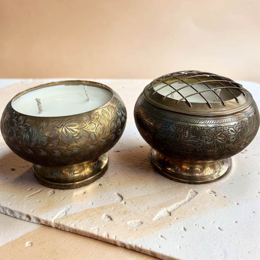 Two ornate brass incense burners, one containing a white candle and the other open with incense material inside, displayed on a textured stone slab against a warm beige background.